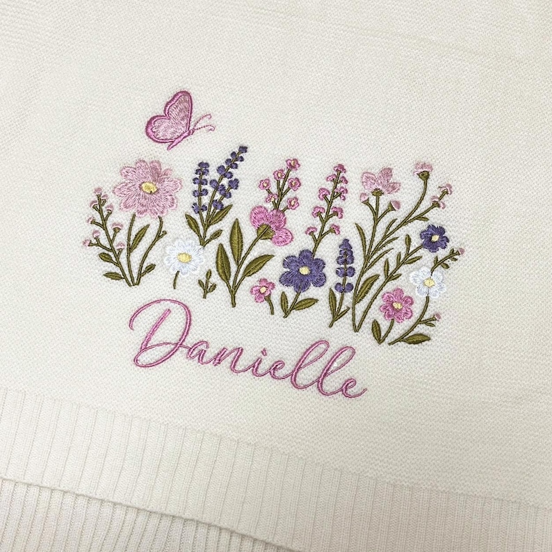 May include: An off-white textile featuring an embroidered design of various wildflowers in shades of pink, purple, and white, accompanied by a small pink butterfly. The name Danielle is embroidered in a cursive pink script below the flowers.