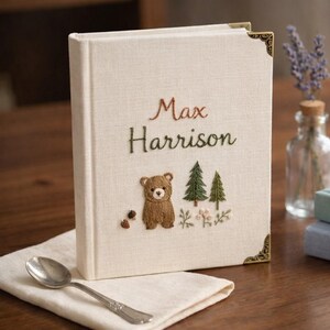 May include: A cream-colored fabric-covered book with gold-tone metal corner protectors. The cover features embroidered text reading Max Harrison above a brown bear, two green pine trees, and small floral accents in brown and green thread.