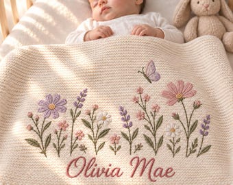 May include: A cream-coloured knit baby blanket featuring embroidered wildflowers in shades of pink, purple, and white, a small purple butterfly, and the name Olivia Mae stitched in pink cursive script across the bottom section of the fabric.