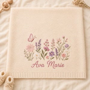 May include: A cream-coloured knit blanket featuring an embroidered design of various wildflowers in shades of pink, purple, and green, accompanied by a small butterfly. The name Ava Marie is embroidered in pink script below the floral arrangement.