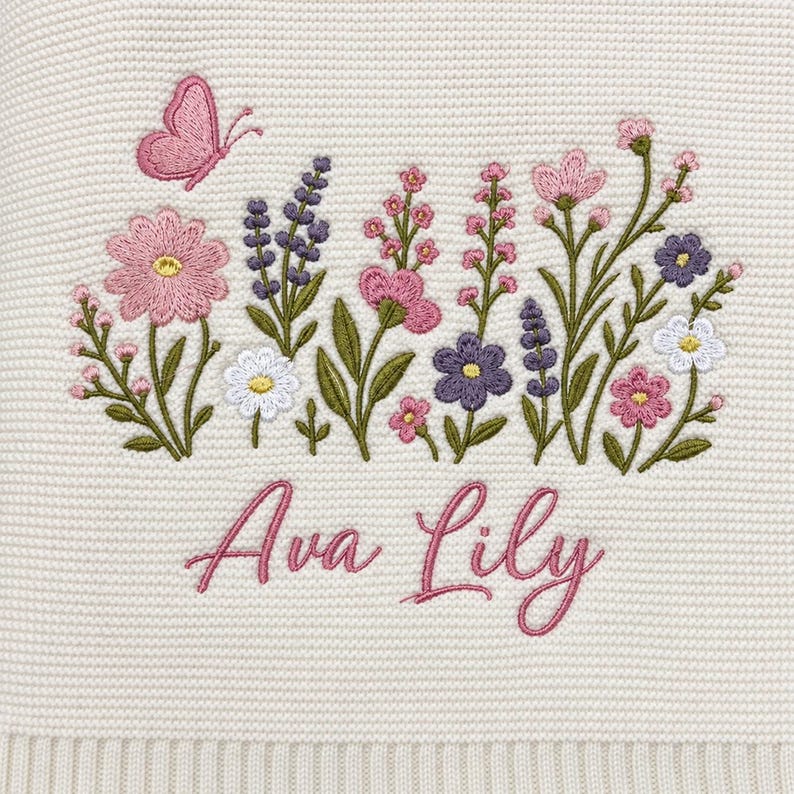 May include: A cream-coloured knit fabric featuring an embroidered design of a pink butterfly and various wildflowers in shades of pink, purple, and white. Below the floral arrangement, the name Ava Lily is embroidered in a pink cursive script.
