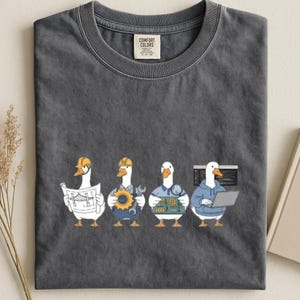 May include: A charcoal gray crew neck t-shirt featuring a graphic of four white ducks in different roles: an engineer with a bridge blueprint, a mechanic with a gear and wrench, a technician with a circuit board, and a programmer with a laptop.