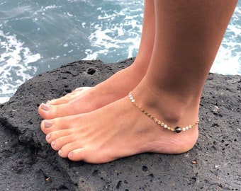 May include: A gold-colored anklet with small, round charms and a dark bead is worn on an ankle. The person is standing on a dark, textured rock near the ocean. The water is a light blue color.