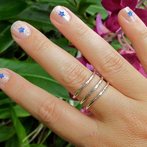 May include: A hand wearing four silver hammered stacking rings. The nails are painted a light silver color with a blue star on each nail.