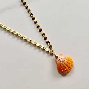 Puede incluir: Un collar de tono dorado con una cadena de eslabones circulares planos y un colgante único que consiste en una pequeña concha marina de color naranja y rosa envuelta en un fino alambre de color dorado que cuelga de la cadena.