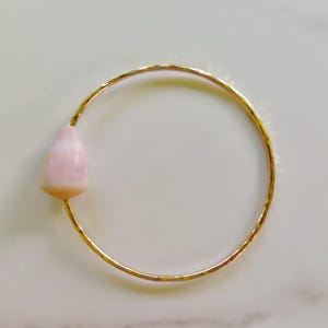 May include: A thin, circular gold-toned bangle bracelet with a hammered texture. A single, smooth, teardrop-shaped light pink shell bead is attached to one side of the metal band, creating a simple and distinct focal point on the jewellery piece.