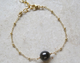May include: A delicate gold-toned bracelet featuring a single, large, dark gray pearl. The bracelet has a fine chain design with small, evenly spaced gold beads. The clasp is adjustable.