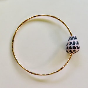 May include: A thin, circular gold bangle bracelet with a hammered texture. A single, small, cone-shaped seashell with a light pink base and dark brown, chevron-like geometric markings is attached to the side of the gold metal band.