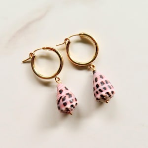 May include: A pair of gold hoop earrings, each featuring a dangling, cone-shaped seashell charm. The shells are light pink with dark brown, rectangular-shaped spots distributed across their surfaces. The hoops are thin, circular, and metallic gold.