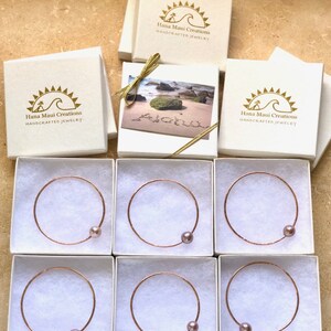 May include: Six white boxes with gold lettering that reads "Hana Maui Creations HANDCRAFTED JEWELRY". Each box contains a rose gold bangle with a single, light purple pearl.