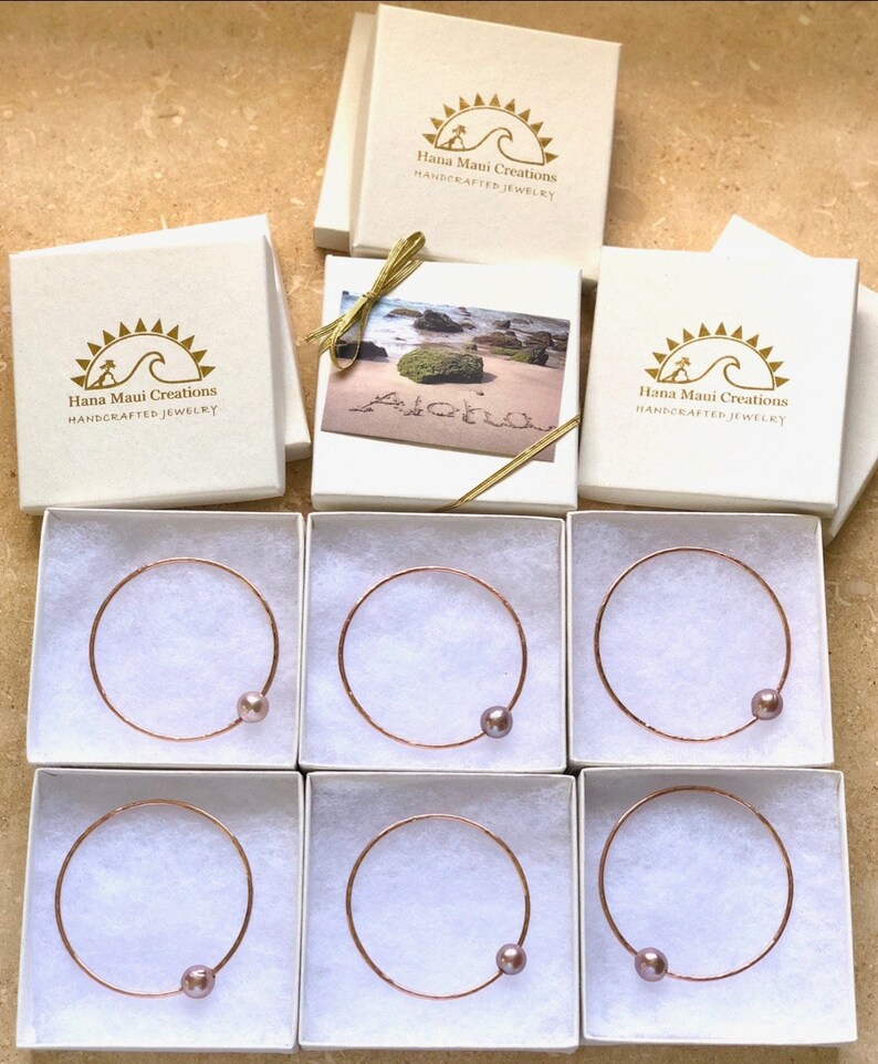 May include: Six white boxes with gold lettering that reads "Hana Maui Creations HANDCRAFTED JEWELRY". Each box contains a rose gold bangle with a single, light purple pearl.