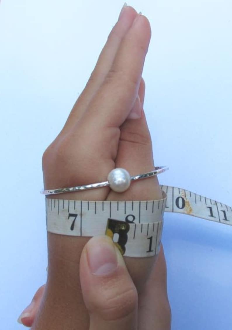 May include: A silver bangle bracelet with a white pearl charm. The bracelet is being measured with a tape measure, showing a measurement of 7 inches.