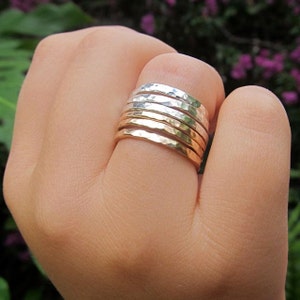 Könnte beinhalten: Ein Satz von sieben gehämmerten Stapelringen in Silber- und Goldtönen. Die Ringe werden an einem Finger getragen.