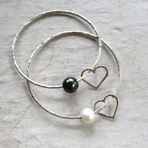May include: Two silver bangle bracelets with hammered texture, one with a black pearl and a cutout heart charm, the other with a white pearl and a cutout heart charm.