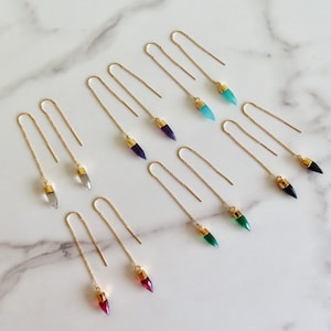 Könnte beinhalten: Eine Sammlung goldfarbener Ohrringe mit farbigen Edelsteinanhängern. Die Ohrringe haben klare, violette, türkisfarbene, schwarze, grüne und rote Edelsteine. Die Edelsteine sind kugelförmig und haben goldene Kappen.