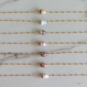Può includere: Sei delicate collane con piccoli pendenti a forma di conchiglia conica. I gusci variano di colore, dal crema e rosa al marrone maculato. Ogni conchiglia è infilata su una sottile catena color oro con minuscoli dettagli di perline, creando un design di gioielli minimalista ispirato alla spiaggia.