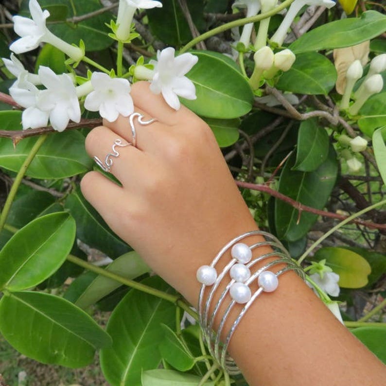 May include: A silver bracelet with multiple strands and white pearls. The bracelet is worn on a wrist with two silver rings, one with the word "love" and the other with a wave design.