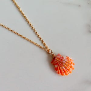 May include: A gold-colored chain necklace with a pendant shaped like an orange and white seashell. The seashell is wrapped with gold-colored wire. The necklace is displayed on a white surface.