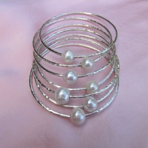 May include: A set of eight silver bangles with a hammered finish, each adorned with a white pearl. The bangles are stacked on top of each other, creating a layered effect.