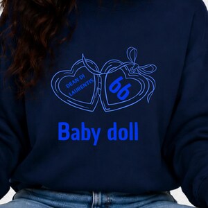 Op de afbeelding: Marineblauw sweatshirt met een blauwe afbeelding van twee harten die met een strik zijn verbonden. De tekst "Dean of Laurentis" en het nummer "66" staan in de harten. Het woord "Baby doll" staat eronder in blauw.