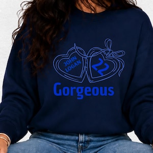 Op de afbeelding: Marineblauw sweatshirt met een blauwe afbeelding van twee hartvormige hangers, een met de naam "John Logan" en de andere met het nummer "22". Het woord "Gorgeous" staat eronder in het blauw.