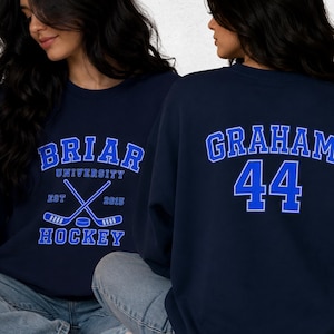 Op de afbeelding: Marineblauw sweatshirt met de tekst "BRIAR UNIVERSITY HOCKEY" in blauw en wit. Het ontwerp aan de voorkant bevat gekruiste hockeysticks en een puck. De achterkant toont "GRAHAM 44" in blauw en wit.