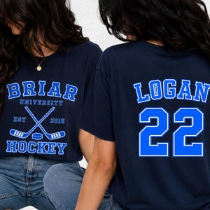 Pode incluir: Camiseta azul marinho com o texto "BRIAR UNIVERSITY HOCKEY" em branco e azul. A frente da camisa apresenta tacos de hóquei cruzados e um disco. A parte de trás da camisa exibe o nome "LOGAN" e o número "22" em azul.