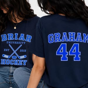 Op de afbeelding: Marineblauwe t-shirts met witte en blauwe tekst. Eén shirt zegt "Briar University Hockey" met gekruiste hockeysticks en een puck. Het andere shirt heeft "Graham" en het nummer "44" op de rug.