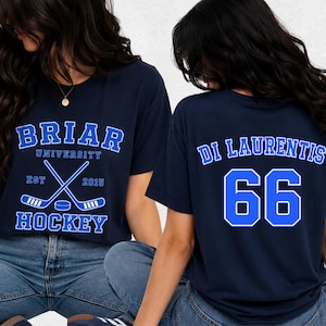 Op de afbeelding: Marineblauwe T-shirts met witte en blauwe tekst. Eén shirt zegt "Briar University Hockey" met gekruiste hockeysticks. Het andere shirt heeft "Di Laurentis" en het nummer "66" op de rug. Beide shirts worden door een persoon gedragen.
