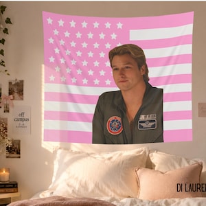 May include: A pink and white American flag tapestry with a portrait of a man in a green flight suit. The tapestry is hanging on a white wall, with other decorative items.