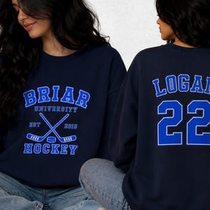 Op de afbeelding: Marineblauw sweatshirt met de tekst "BRIAR UNIVERSITY HOCKEY" in blauw en wit. Het ontwerp bevat gekruiste hockeysticks en een puck. De achterkant toont "LOGAN 22" in blauw en wit.