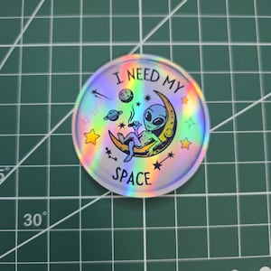 Puede incluir: Una pegatina holográfica circular con el texto "I NEED MY SPACE". Presenta un alienígena de dibujos animados descansando en una luna creciente, con estrellas, planetas y un efecto arcoíris.