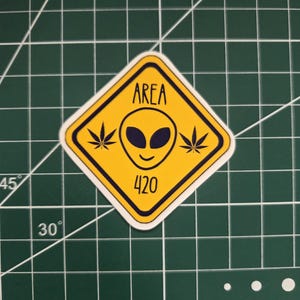 May include: A diamond-shaped yellow sticker with a black border featuring a central alien head graphic, two cannabis leaf icons on either side, and the black text "AREA" above and "420" below the alien head, set against a green cutting mat.