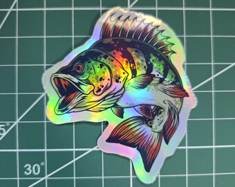 May include: Holographic sticker of a fish with an open mouth, featuring a rainbow of colors including green, yellow, and red. The sticker is on a green cutting mat with grid lines and degree markings.