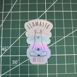 Op de afbeelding: Een holografische sticker met een lama in een meditatieve pose. De sticker heeft de tekst "LLAMASTE" boven de lama en "IN BED" eronder. De lama is in tinten blauw, paars en roze.