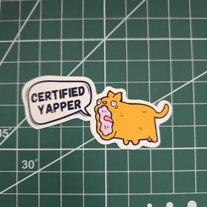 Op de afbeelding: Een gestanste sticker met een oranje cartoonkat met de mond wijd open in een blaffende uitdrukking, naast een witte tekstballon met de donkerblauwe tekst "CERTIFIED YAPPER" in een vetgedrukt, schreefloos lettertype.