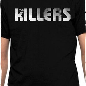 Op de afbeelding: Zwart T-shirt met de bandnaam "the Killers" in een witte, gepixelde lettertype. Het shirt met korte mouwen is gemaakt van een donkere stof en wordt door een persoon gedragen.