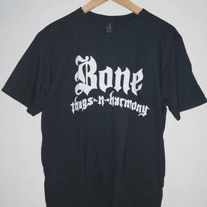 May include: Black t-shirt with the white text "Bone thugs-n-harmony" in a gothic font. The shirt is hanging on a wooden hanger against a white background.