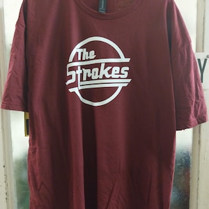 Peut inclure: Un t-shirt à manches courtes bordeaux avec un motif circulaire blanc sur la poitrine. Le motif contient le texte "The Strokes" dans une police stylisée, enfermé dans un cercle blanc avec des lignes horizontales traversant le centre.