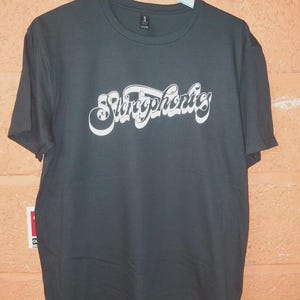 Op de afbeelding: Donkergrijs t-shirt met het woord "Stereophonics" in witte, gestileerde letters op de borst. Het t-shirt hangt aan een witte hanger. De stof lijkt zacht te zijn.