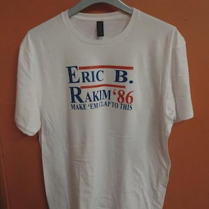 Puede incluir: Camiseta blanca con el texto "Eric B. Rakim '86" en letras azules y rojas. La camiseta también incluye la frase "Make 'em clap to this". La camiseta está colgada en una percha.