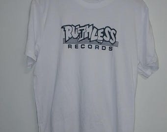 May include: White t-shirt featuring the text "RUTHLESS RECORDS" in dark blue. The design is printed across the chest. The t-shirt is hanging on a wooden hanger against a white wall. It has a crew neck and short sleeves.