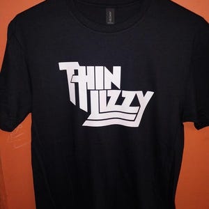 Op de afbeelding: Zwart T-shirt met de bandnaam "Thin Lizzy" in witte blokletters. De tekst is gestileerd met een uniek lettertype. Het T-shirt hangt aan een hanger tegen een oranje achtergrond.