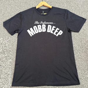 Könnte beinhalten: Schwarzes T-Shirt mit dem weißen Schriftzug "The Infamous... MOBB DEEP" in Bogenform. Das T-Shirt hängt an einem Holzkleiderbügel.