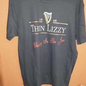 Op de afbeelding: Donkergrijs t-shirt met de tekst "Thin Lizzy", een harp logo en de jaartallen "1969" en "1969". Onder de bandnaam staat de tekst "Whisky In The Jar" in cursief. Het t-shirt hangt aan een witte hanger.