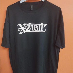 Puede incluir: Camiseta negra con la palabra "Xzibit" en blanco en una fuente estilizada. La camiseta está colgada de una percha blanca sobre un fondo naranja. La camiseta es de cuello redondo.