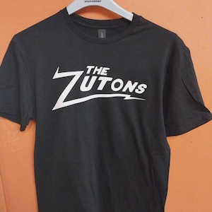 Op de afbeelding: Zwart T-shirt met de witte tekst "THE ZUTONS" in een gestileerd lettertype. Het T-shirt hangt aan een witte hanger met de merknaam "wynnster". De achtergrond is effen oranje van kleur.