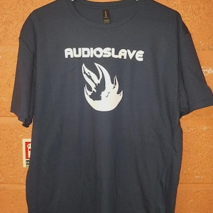 Op de afbeelding: Marineblauw T-shirt met korte mouwen met het woord "AUDIOSLAVE" in witte blokletters boven een witte vlamvormige afbeelding. Het T-shirt hangt aan een grijze hanger.