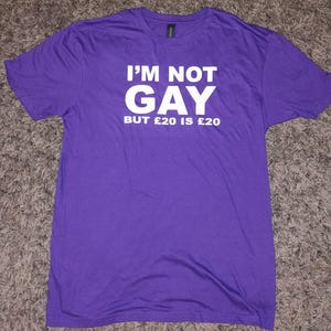 Puede incluir: Una camiseta morada con el texto "I'M NOT GAY BUT £20 IS £20" impreso en blanco. La camiseta es de manga corta y cuello redondo.