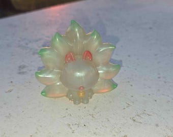 Peut inclure: Figurine nacrée d'un animal stylisé avec un motif floral. La figurine a un corps rose clair, des oreilles rouges et des pétales à bout vert. Un petit charme doré pend de son cou.